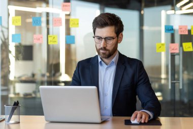 Modern ofis ortamında dizüstü bilgisayara odaklanan işadamı, cam duvardaki renkli yapışkan notlarla çevrili. İş ortamında odaklanma, üretkenlik ve analiz portreleri.