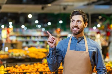 Önlüklü market çalışanı üretim bölümüne işaret ederken gülümsüyor. Canlı meyve ve sebzeler etrafında davetkar, taze bir atmosfer yaratır..