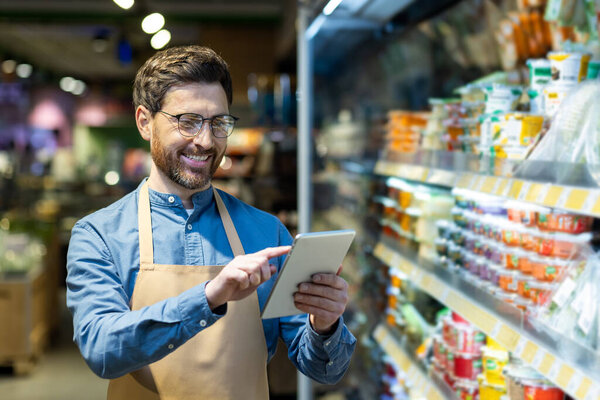 Confident grocery store worker uses digital tablet while standing in supermarket aisle. Surrounded by various products, he engages with technology to manage inventory.