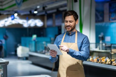 Önlüklü bir süpermarket çalışanı envanter kontrolü için dijital tablet kullanıyor. İmaj, perakende, verimlilik ve modern market ortamındaki teknolojiyi temsil ediyor. Taze ürün ve canlı ortam.