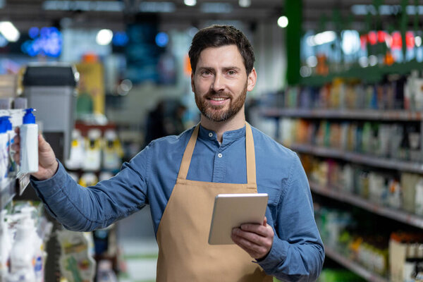 Smiling shopkeeper wearing apron holding tablet in supermarket aisle. Man updates inventory and provides excellent customer service. Retail worker efficiently manages store products and interacts
