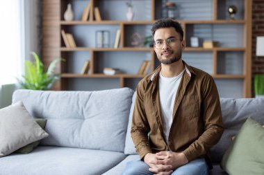 Evdeki kanepede rahat giysiler içinde oturan ve kameraya bakan ciddi bir Hintli adamın portresi..