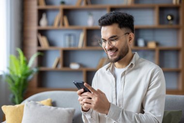 Genç, gülümseyen Hintli bir adamın kanepede oturduğu ve elinde cep telefonu kullandığı fotoğraf..