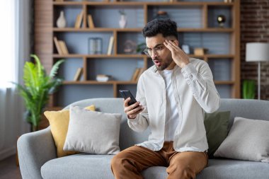 Kafası karışmış genç Hintli adam evde, koltukta oturuyor, cep telefonu ekranına bakıyor ve eliyle kafasını tutuyor..