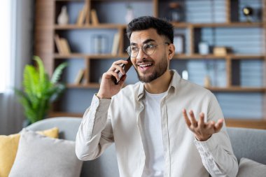 Hintli genç adam gözlüklü ve günlük kıyafetleriyle evdeki kanepede oturuyor ve elleriyle işaret ederken gülümsüyor..
