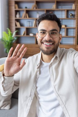 Evde genç bir Hintlinin portresi, elinde kamerayla konuşuyor, el sallıyor ve selamlıyor. Yakın plan dikey fotoğraf.