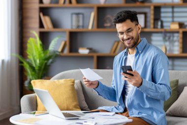 Gülümseyen Hintli genç adam evdeki koltukta oturuyor, elinde belgeler ve faturalar var ve cep telefonuna bakıyor..