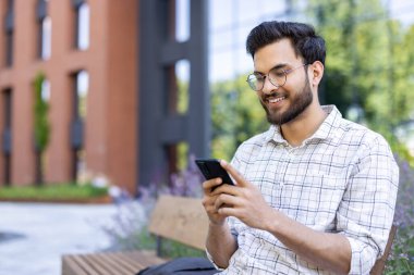 Gülümseyen genç Hintli bir adam dışarıda bankta oturuyor ve cep telefonu kullanıyor..