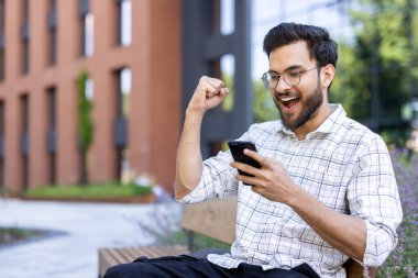 Şehirdeki bankta oturan gözlüklü genç adam telefon kullanırken heyecanını dile getiriyor. Başarı, mutluluk, başarı ve teknoloji kavramı.