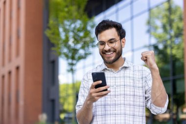 Gözlüklü ve ekose gömlekli genç adam dışarıda telefona bakarken başarıyı kutluyor. Yumruğunu sıkmış neşeli bir adam binanın ve cam pencerelerin yanında durmuş, sevinçlerini ifade ediyor.