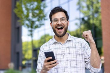 Sokakta elinde cep telefonuyla bekleyen ve kameranın zaferini kutlayan gülümseyen genç bir Hintlinin yakın plan portresi..