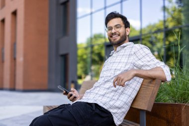 Genç Hintli bir adam dışarıda bir bankta, elinde telefonla rahatça oturuyor. Gülümseyerek kameraya bakıyor..