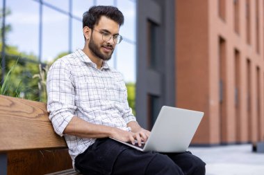 Genç adam dizüstü bilgisayar kullanarak bankta oturuyor. Modern binanın yakınındaki açık hava ortamında çalışmak ya da ders çalışmak. Sıcak ve verimli bir atmosfer..