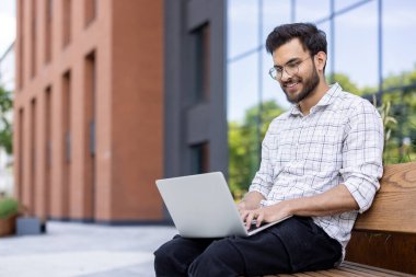 Şehirdeki bankta otururken dizüstü bilgisayar kullanan gülümseyen adam. Gözlüklü ve rahat gömlekli bir adam uzaktan çalışıyor. Modern teknoloji, uzaktan çalışma, açık hava verimliliği..
