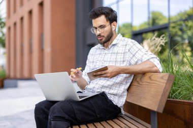 Genç adam kulaklık takarken dizüstü bilgisayar ve telefon kullanarak bankta oturuyor. Arka planda yansıtıcı modern ofis binası, günlük iş yeri sahnesi