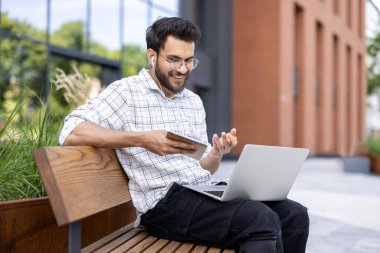Hintli gülümseyen bir adam sokakta bir bankta oturmuş elinde kalemle bir not defteri tutarak bilgisayarla video konuşuyordu..