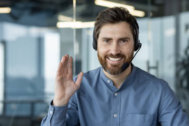 Ofiste kulaklık takmış, kameranın önünde oturmuş gülümseyen, selamlayan ve el sallayan genç bir adamın yakın plan portresi..