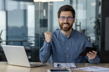 Gözlüklü bir gencin portresi masada oturuyor ve finansal başarının tadını çıkarıyor, elinde bir hesap makinesi tutuyor, eliyle evet işareti yapıyor, kameraya bakıyor..