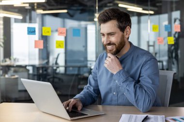 Gülümseyen genç bir iş adamı ofiste dizüstü bilgisayarla çalışıyor, masada oturuyor ve ekrana bakıyor..