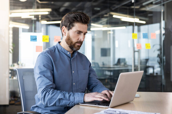 Молодой серьезный человек работает по очереди за столом в офисе, сидя и печатая на клавиатуре ноутбука.