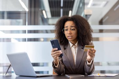 Worried young african american woman in suit sitting at desk in office, holding credit card and looking upset at mobile phone. clipart