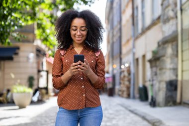 Genç bir Afro-Amerikalı kadın sırt çantasıyla sokakta duruyor ve gülümsüyor cep telefonu kullanıyor, mesaj yazıyor, bilgi arıyor..