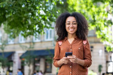 Sırt çantalı, gözlüklü, şehir sokağında elinde telefonla kameraya gülümseyen Afro-Amerikan bir kadının portresi..