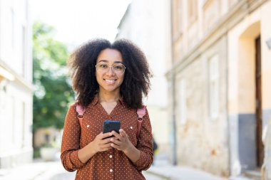 Sokakta elinde telefonla kameraya bakıp gülümseyen kıvırcık saçlı Amerikalı bir kızın portresi..