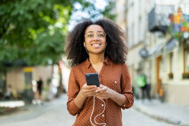 Kulaklıkla gülümseyen Afro-Amerikalı bir kız, elinde telefon, şehirde yürüyor, müzik dinliyor ve konuşuyor..