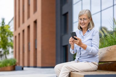 Kentteki akıllı telefonlara bakarak bankta oturan gülümseyen son sınıf öğrencisi. Günlük giysiler. Teknolojiyle uğraşırken açık havada rahatlama.