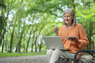 Diz üstü bilgisayar ve not defteri kullanarak sakin bir ortamda internetten öğrenen gülümseyen bir kadın. Bilgiyi ve doğayı yakalayarak, üretken öğrenme için teknoloji ve sükunetin sinerjisinden zevk alıyor..