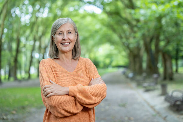 Зрелая женщина с седыми волосами и оранжевым свитером уверенно стоит в пышном зеленом парке. Она тепло улыбается, символизируя счастье, спокойствие и признательность природе..