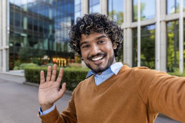 Modern ofis binasının dışında selfie çeken neşeli bir adam. İnsanlar gülümsüyor, el sallıyor, misafirperver bir ortam yaratıyor. Parlak güneşli bir gün, olumlu ve profesyonel havayı arttırır..