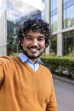 Gülümseyen adam modern ofis binasının dışında selfie çekiyor. Süveter ve gömlekle kendine güvenini ve günlük tarzını gösterir. Güneşli bir günde açık şehir ortamını sever, neşeli bir görüntü yakalar.