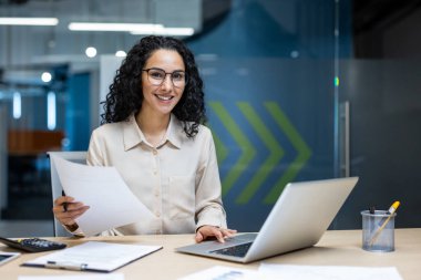 Modern ofis masasında oturan kendine güvenen bir iş kadını. Bilgisayarda evrak işleri ve hesap makinesiyle çalışıyor. Dostça atmosferli profesyonel ortam, verimlilik ve kariyer başarısını gösteriyor.