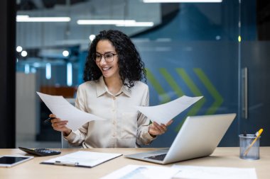 İş kadını büro masasında oturmuş belgeleri inceliyor. Gülümseyen profesyoneller bilgisayar ve telefonla birden fazla iş yapıyor, yaratıcı çalışma alanındaki verimliliği gösteriyor.