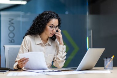 İş kadını telefonda birden fazla iş yaparken ve kağıt belgelerini incelerken ciddi evrak işlerine odaklandı. Modern ofiste masa başında oturan kadın, iş iletişimi için dizüstü bilgisayar kullanıyor..