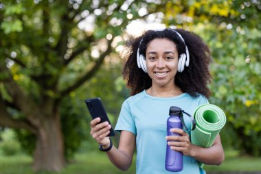 Dışarıda spor salonunda yoga minderi ve su şişesi tutan, kulaklıkla müzik dinlerken akıllı telefon kullanan gülümseyen bir kadın. Doğal ortamdaki aktif yaşam tarzının tadını çıkarıyorum.