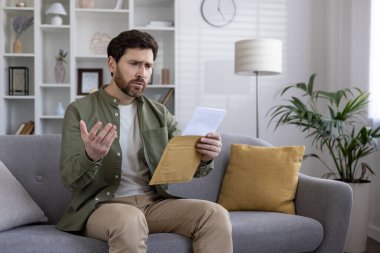 Kanepede sakallı bir adam kafası karışmış bir ifadeyle mektuba bakıyor. İçeriği hakkında endişeli ve kararsız görünüyor. Modern oturma odasının ayarlanması ev içi stresi arttırır.