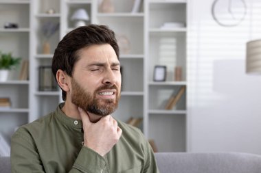 Boğazı ağrılı bir ifadeyle tutulan bir adam boğaz ağrısı ya da rahatsızlık hissi veriyor. Görüntü, muhtemelen grip, soğuk algınlığı ya da alerjiyle ilgili sağlık sorunları yaşayan yetişkin bir erkeğin resmini çekiyor.