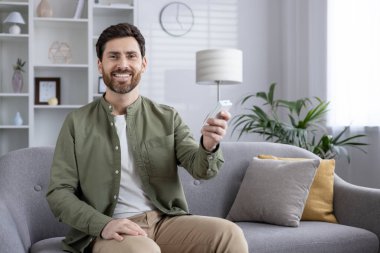 Çağdaş oturma odasındaki koltukta oturan gülümseyen adam elinde uzaktan kumandayla klima tutuyordu. Günlük hayatta rahatlık, rahatlama ve teknolojiyi yakalar..