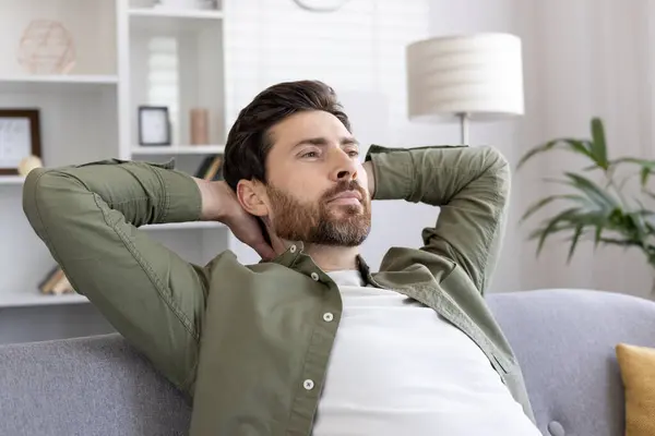 stock image Relaxed man sits on couch with hands behind head, reflecting in modern living room. Casual attire complements calm demeanor, conveying tranquility and peacefulness. Relaxation and contemplation