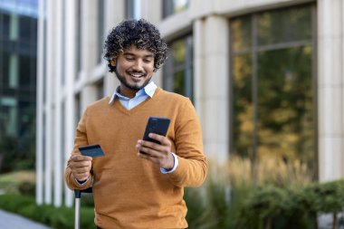 Gülümseyen adam elinde telefon ve kredi kartıyla modern binanın önünde duruyor. Günlük süveter giyiyor, online alışveriş veya dijital bankacılık ile ilgileniyor gibi görünüyor, teknoloji ve kolaylığı birleştiriyor.