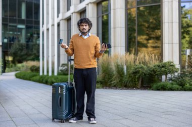 Ofis binasının dışında kafası karışmış, elinde akıllı telefon ve kredi kartı tutan bir adam var. Çanta taşıyor, muhtemelen seyahat ya da ödeme sorunu yaşıyor. Kredi kartı dolandırıcılığı veya seyahat kavramı
