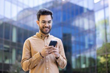 Genç adam gülümserken modern cam ofis binasının dışında akıllı telefon kullanıyor, şehir yaşam tarzını, bağlantıyı ve iletişimi sembolize ediyor.