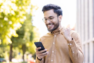 Genç adam akıllı telefon kullanarak dışarıda başarıyı kutluyor. Mutlu ve sıradan kıyafetlerle heyecanlı görünüyor, bir dönüm noktası yaşıyor. Başarı kavramları ve neşeli haberler için idealdir..