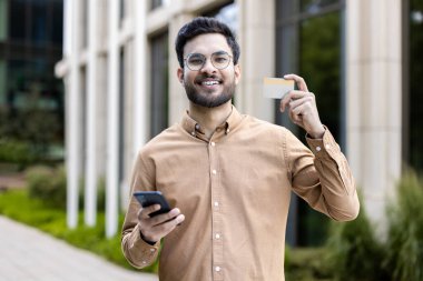 Bir elinde kredi kartıyla dışarıda duran genç bir adam diğer elinde akıllı bir telefon, güvenle gülümsüyor. Görüntü modern teknoloji kullanımı, kişisel finans ve dijital yaşam tarzını temsil etmektedir..