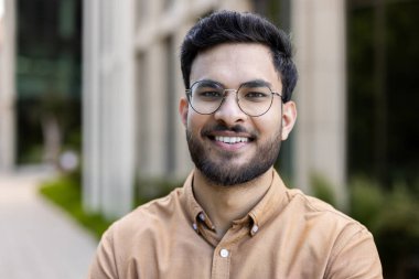 Gözlüklü ve sakallı genç bir adam açık hava ortamında güvenle gülümsüyor. Sıradan bir gömlek giyiyor, cana yakın ve memnun görünüyor. Rahatlama ve olumlu zihniyet tasvir eder.