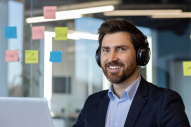 Kulaklık takan gülümseyen iş adamı ofis masasında çalışıyor. Etrafı renkli yapışkan notlarla çevrili bilgisayar görevlerine odaklanmış. Modern işyeri, verimlilik ve profesyonel konsantrasyonu temsil eder.