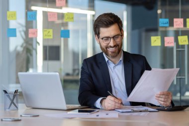 Gözlüklü ve sakallı bir işadamı büro masasında oturmuş elinde kalemle belgeleri inceliyor. Dizüstü bilgisayarla çevrili, pencere notları, verimlilik ve odaklanma hissi.